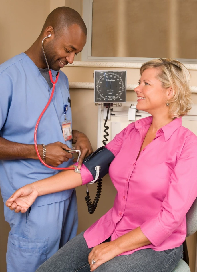 A healthcare worker in blue scrubs checking a woman's blood pressure in a clinic